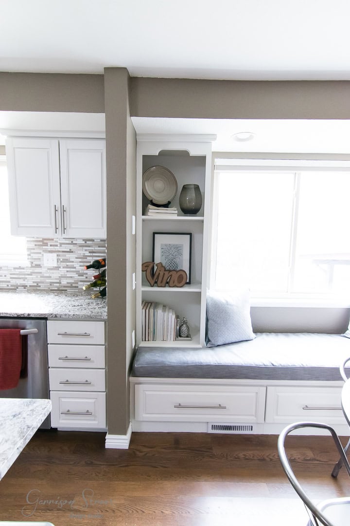 A bright kitchen with white cabinets and a built-in window seat with gray cushions. Shelves above hold decorative items, including a sign reading "Vino." A window lets in natural light, illuminating the wooden floor and neutral walls.