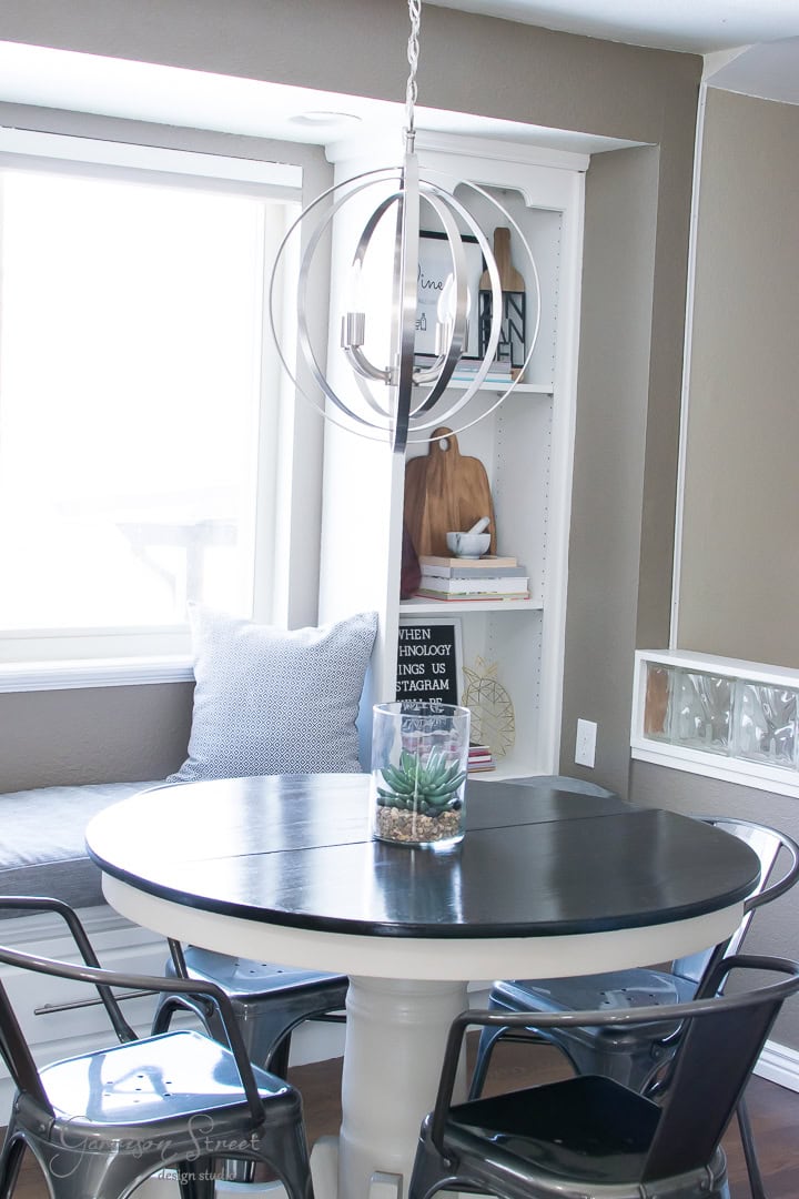A cozy dining nook features a round black table and metal chairs. A modern chandelier hangs above, and a window seat with gray cushions is on the side. Shelves in the background hold decorative items like wooden boards and framed pictures.