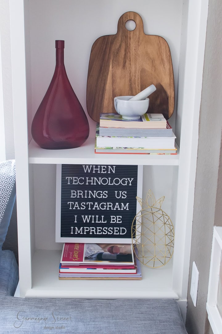 A white shelf holds a maroon vase, a wooden cutting board, a mortar and pestle, and stacked books. A letter board reads, "When technology brings us tastagram I will be impressed." A wire pineapple decor and more books are on the lower shelf.