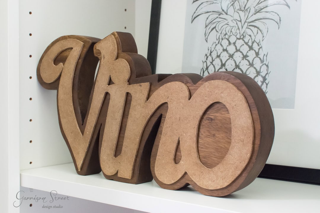 Wooden decor piece displaying the word "Vino" in stylized letters on a white shelf. Behind it is a framed black and white pineapple illustration.