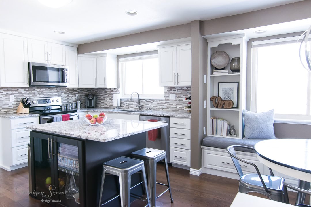 A modern kitchen with white cabinets, a black island with stools, and stainless steel appliances. A cozy window seat is adorned with cushions and decorative items. The room is bright with natural light from large windows.