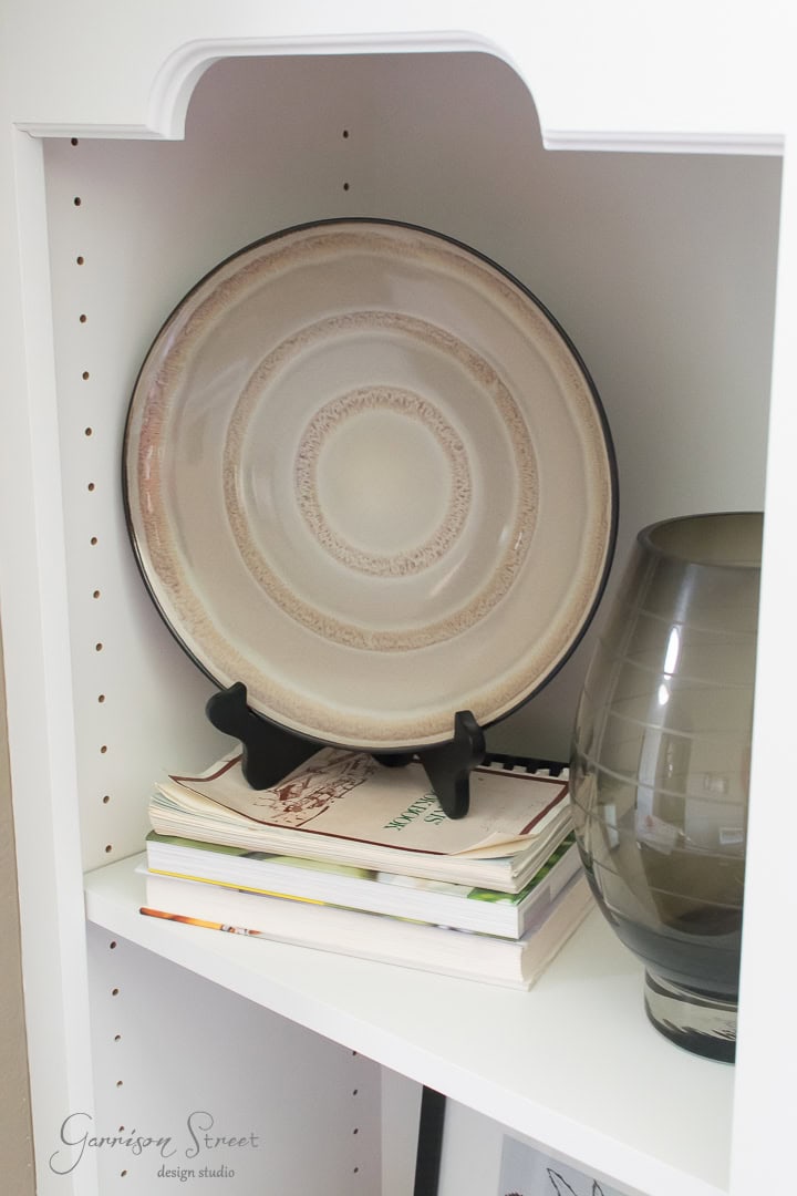 A decorative plate with concentric circle patterns is displayed on a black stand atop a stack of books on a white shelf. Next to it is a translucent gray vase. The shelf has an arched top design.