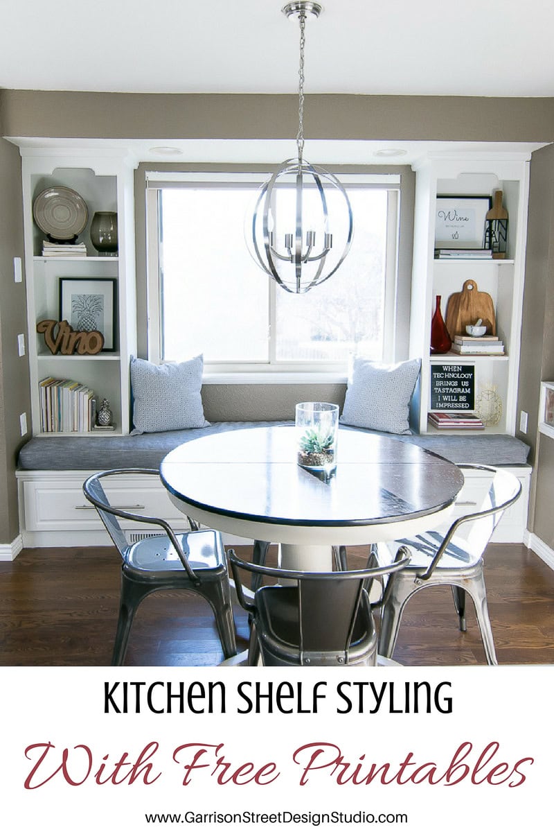 A cozy dining nook with a round table and metal chairs, surrounded by built-in shelves and a cushioned bench. Decor includes framed art, books, and decorative items. A modern chandelier hangs above, and large windows let in natural light.