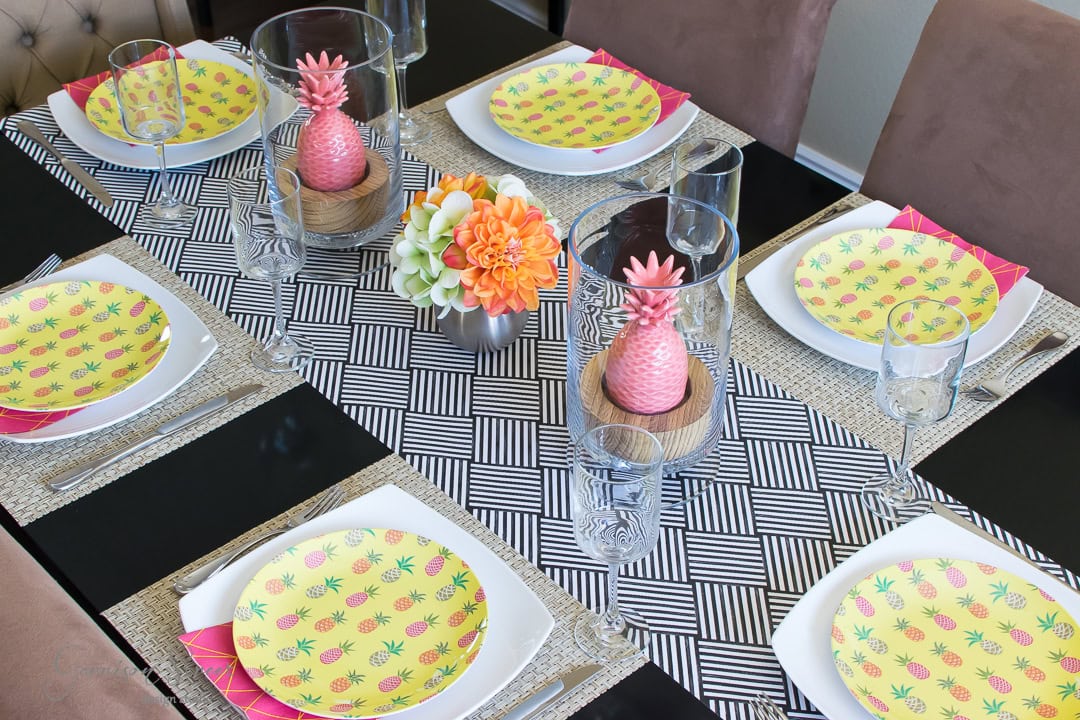 A dining table set for six, featuring colorful plates with pineapple patterns, pink pineapple ornaments under glass cloches, and a floral centerpiece with orange and white flowers. Alternating geometric and woven texture placemats.
