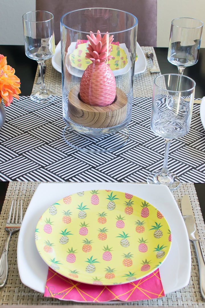 A dining table set with tropical-themed plates featuring colorful pineapples, clear wine glasses, and a pink pineapple ornament in a glass cylinder. The table has a geometric-patterned runner and vibrant orange flowers as a centerpiece.