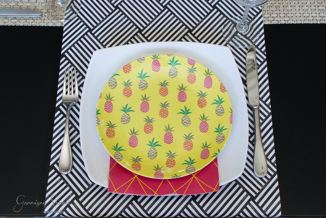 A colorful table setting featuring a yellow plate with pineapple illustrations, placed on a red napkin and white square plate. The background is a black and white geometric placemat, accompanied by a fork, knife, and glass.