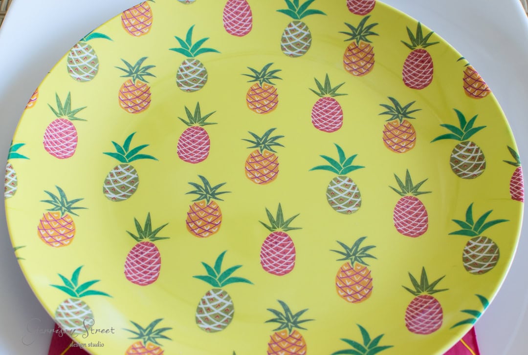 A round yellow plate decorated with repeating patterns of colorful pineapples in pink, green, and white on a textured surface. The plate is placed on a white dish.