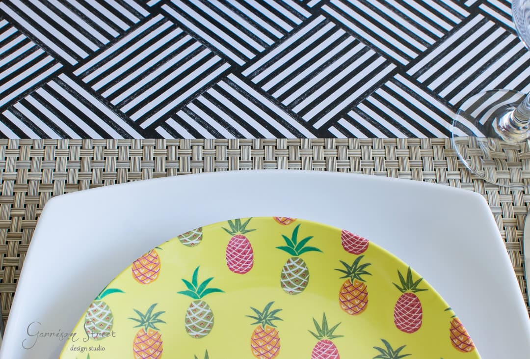 A square white plate with a yellow pineapple-patterned plate on top sits on a wicker-style placemat. Below the plates is a part of a black and white geometric tablecloth. The edge of a wine glass is visible in the top right corner.