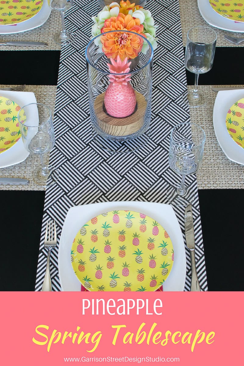 A dining table set with a vibrant pineapple-themed decor, including yellow plates with pineapple patterns and a central floral arrangement in a pink vase. The table features a black and white geometric runner and neatly arranged cutlery and glasses.