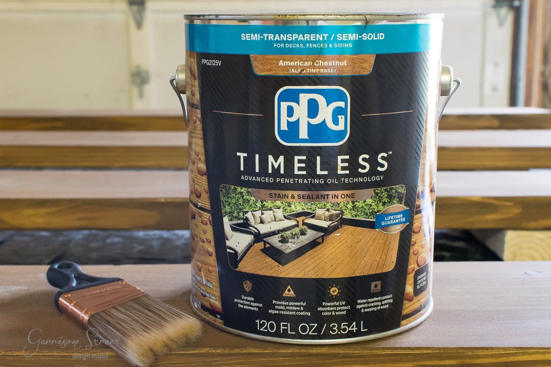 A can of PPG Timeless wood stain in American Chestnut color sits on a wooden surface. A paintbrush is placed beside it. The can highlights features such as semi-transparent finish and advanced penetrating oil technology.
