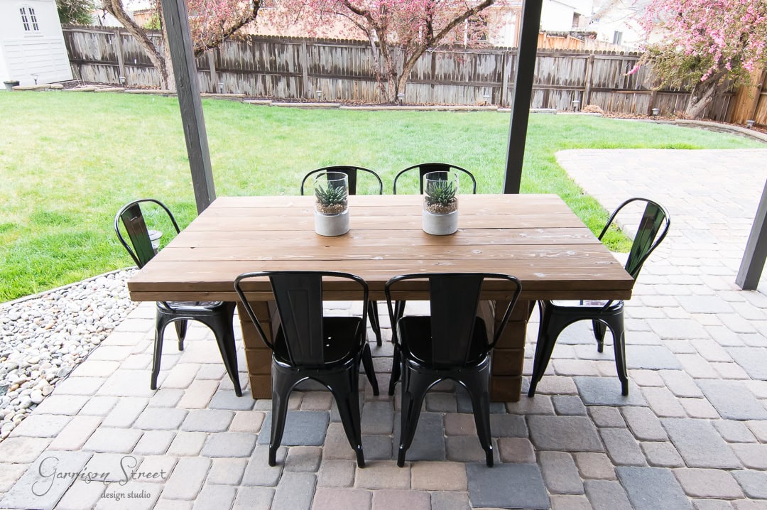 A patio with a wooden dining table and six black metal chairs on a stone floor. Two potted plants sit on the table. The area is surrounded by a grassy yard and wooden fence, with blooming trees in the background.