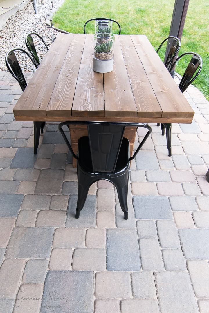 A wooden outdoor table with a succulent centerpiece, surrounded by six black metal chairs, is set on a patio with light and dark gray stone pavers. Grass is visible in the background.