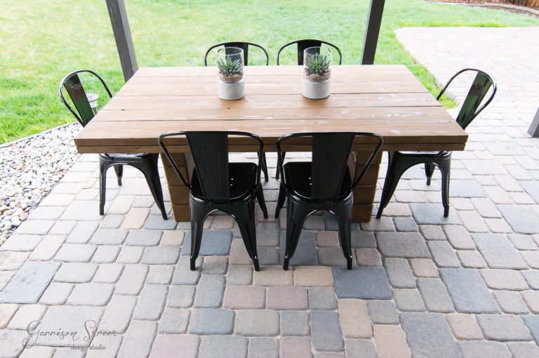 A rectangular wooden table with six black metal chairs is set on a patio with stone pavers. Two small potted plants are placed on the table. A grassy lawn is visible in the background.