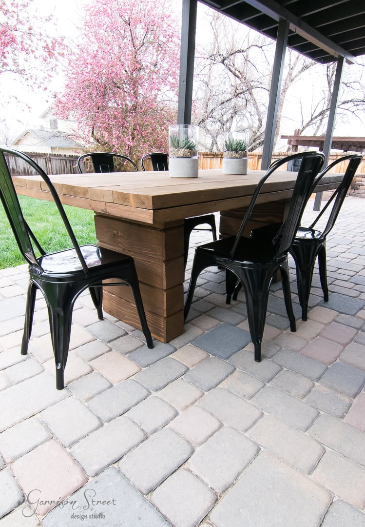 Outdoor patio scene with a wooden table and six black metal chairs on a stone tile floor. Two small potted plants sit on the table. A cherry blossom tree is blooming in the background near a wooden fence.