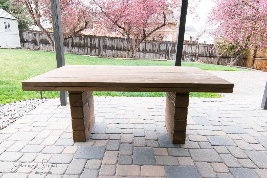 A rectangular wooden table with thick block legs stands on a stone patio. In the background, a grassy yard is bordered by a wooden fence and pink flowering trees.