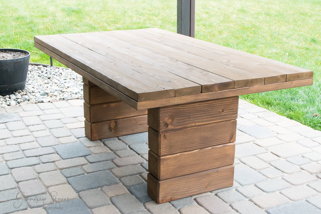Wooden dining table with a rustic design on a stone patio. The table has a sturdy rectangular top and two wide, block-like legs. A green lawn is visible in the background, along with a black planter containing small stones.