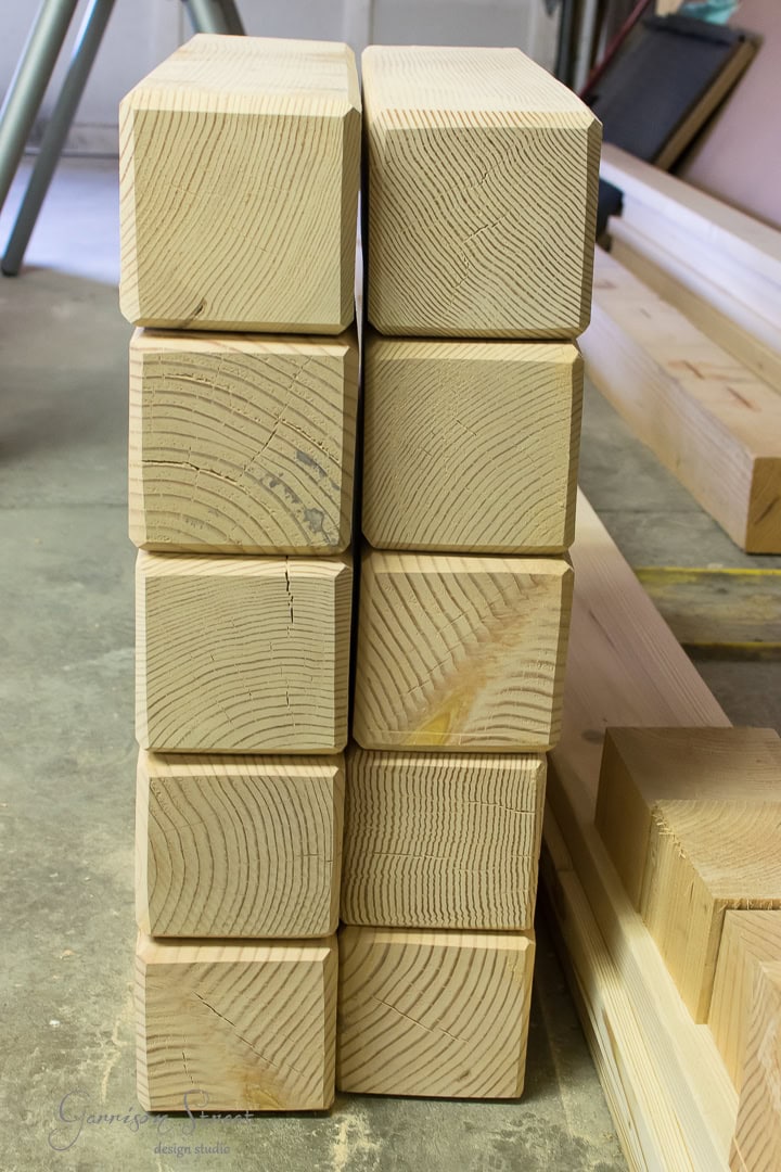 Stacks of neatly arranged, smooth wooden beams with visible grain patterns. The beams are placed upright on a concrete floor in a workshop, alongside other pieces of cut lumber and construction materials.
