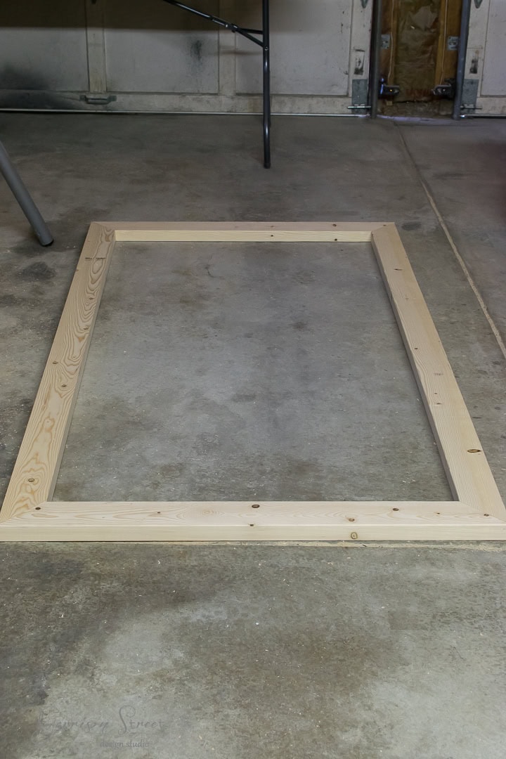 A wooden rectangular frame lies flat on a concrete floor in a garage. The frame is constructed from light-colored wood planks and is assembled with screws. Some tools and materials are visible in the background.
