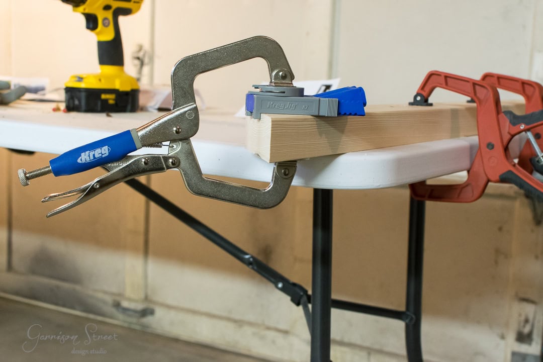 A woodworking clamp secures a piece of wood on a table. Nearby, a drill and another clamp are visible. The setup appears ready for a woodworking project in a workshop setting.