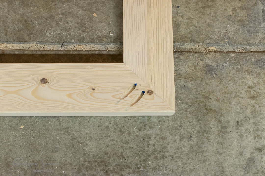 Close-up of two wooden planks joined at a right angle with pocket hole screws. The wood has visible knots and grains, resting on a concrete floor. The screws are inserted into angled holes for a secure connection.