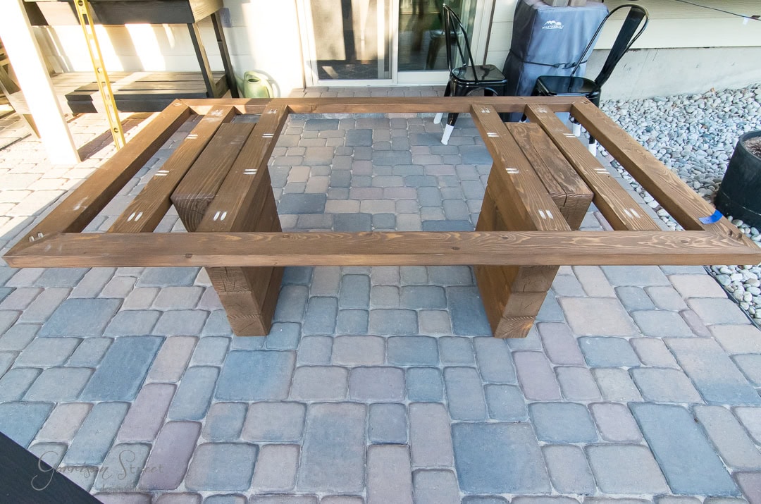 Wooden frame of a rectangular table on a patio with cobblestone flooring. The frame is supported by two wide legs and is situated outdoors near a building with a sliding glass door. A chair and some pebbles are in the background.