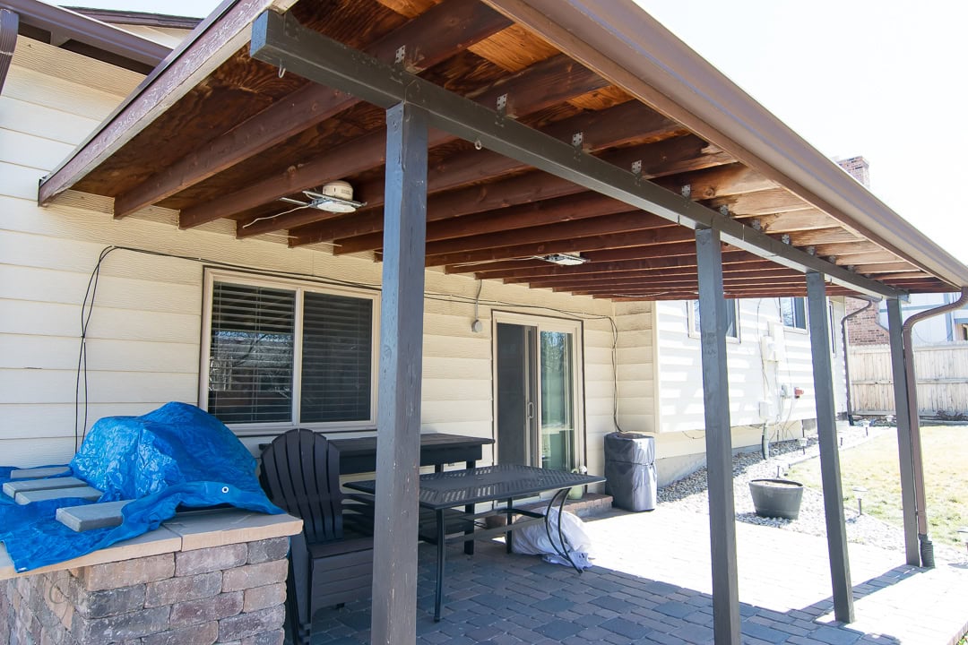 A covered patio with wooden beams and a metal roof extends from a house. There's a table, chairs, and a blue tarp covering an object. The ground is paved with bricks, and a sliding glass door leads inside. A grassy yard is visible in the background.