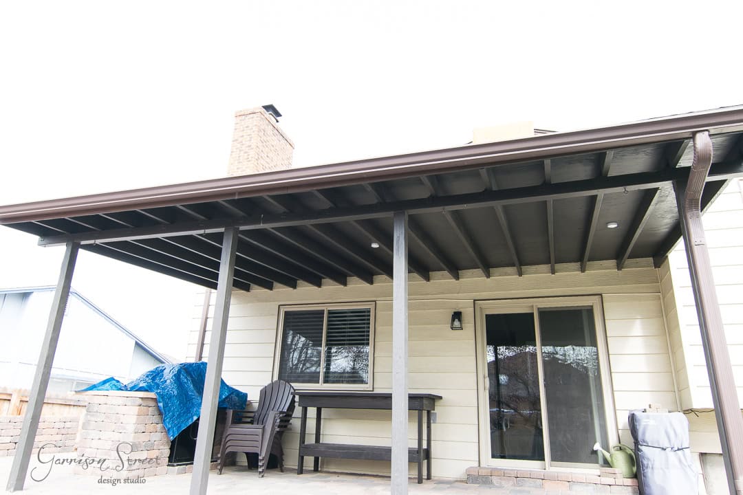 A patio with a dark wooden pergola attached to a house. Underneath are two black chairs and a small table. A blue tarp covers an object to the left, and a chimney is visible on the roof. The house has beige siding and a brick base.