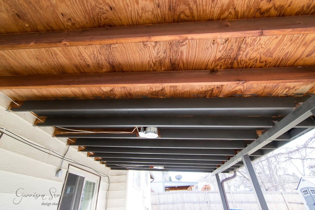 View of a backyard patio showing exposed wooden beams and ceiling with a fan fixture. The structure is supported by vertical metal posts. The surrounding area includes a fence and outdoor elements.