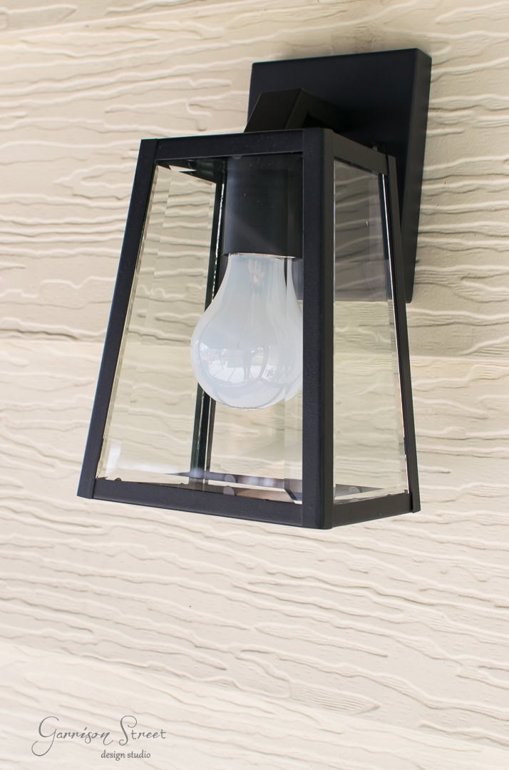A black-framed outdoor wall lantern with a clear glass enclosure, housing a single light bulb, mounted on a textured beige wall.