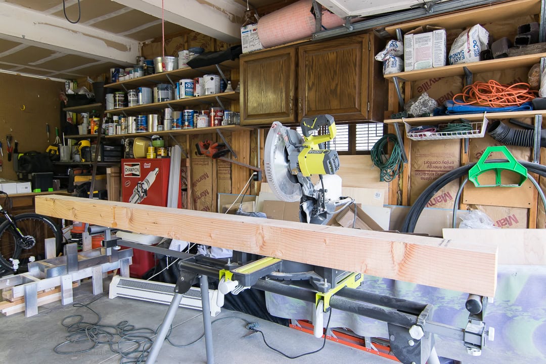 A workshop with a miter saw on a stand, cutting a long wooden beam. Shelves in the background hold paint cans, tools, and other materials. Cables and safety gear are also visible, cluttering the space.