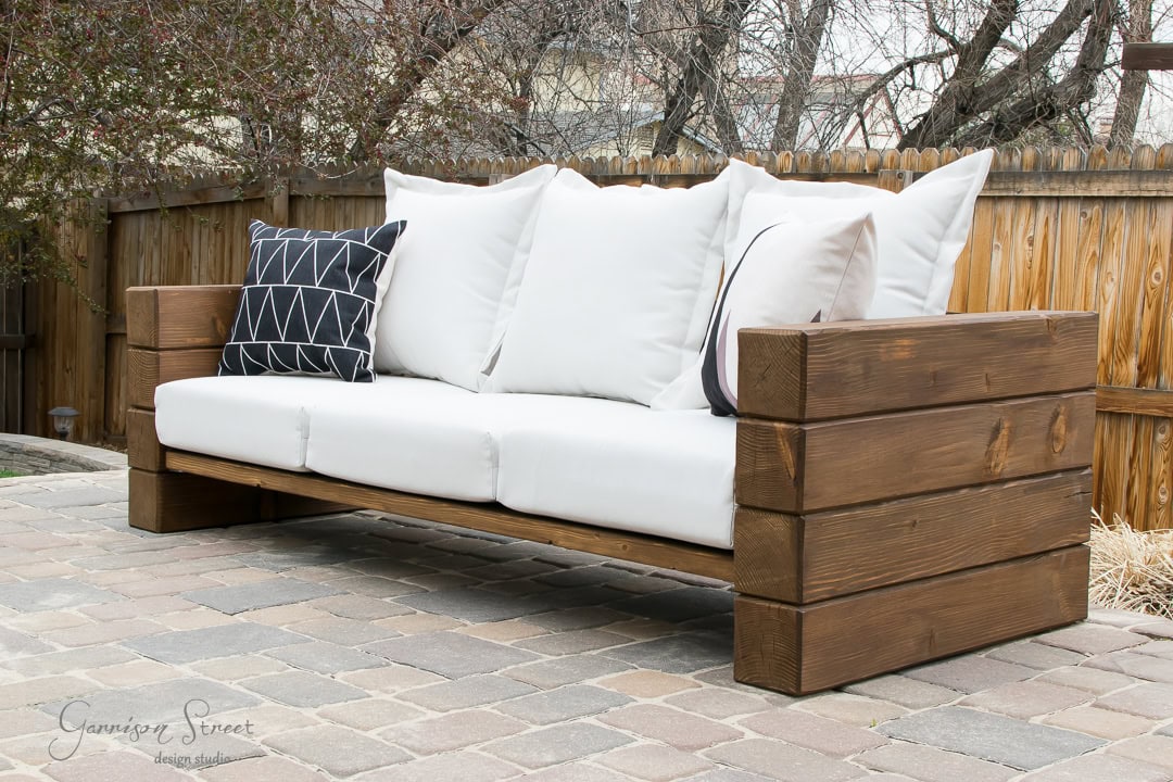 A wooden outdoor sofa with white cushions and two black-and-white patterned throw pillows is placed on a stone patio. A wooden fence and bare trees are visible in the background.