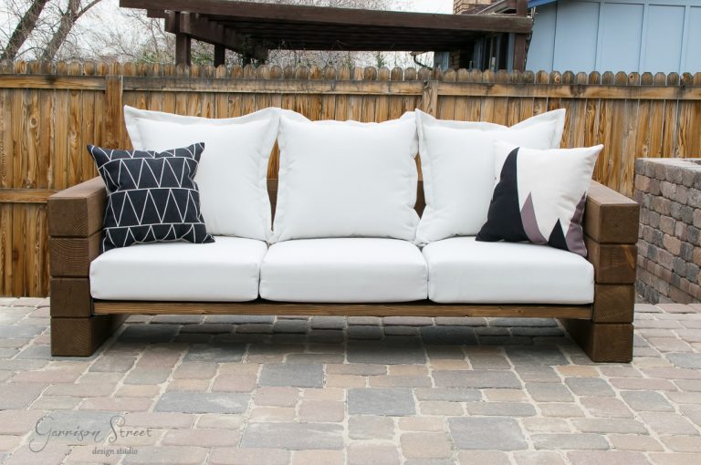 A wooden outdoor sofa with white cushions is set on a stone patio. The sofa has three large white pillows and two decorative pillows, one with a geometric pattern and another with a minimalist design. A wooden fence is in the background.