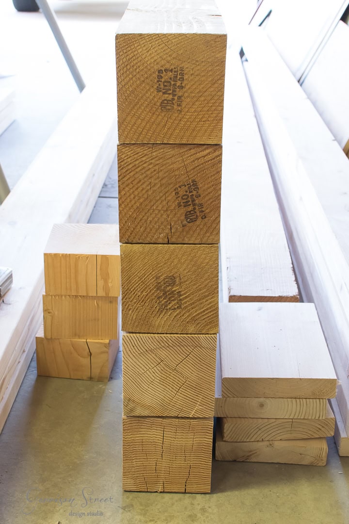 Stack of large, square wooden beams standing on end in a workshop, with smaller wooden pieces beside them. Sunlight casts soft shadows on the concrete floor.