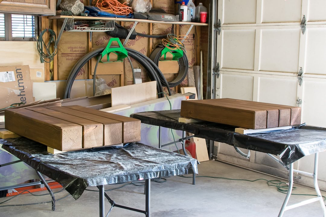 Wooden beams are laid out on two black, tarp-covered tables inside a garage. The space is cluttered with various tools and materials, and the garage door and walls are visible in the background.
