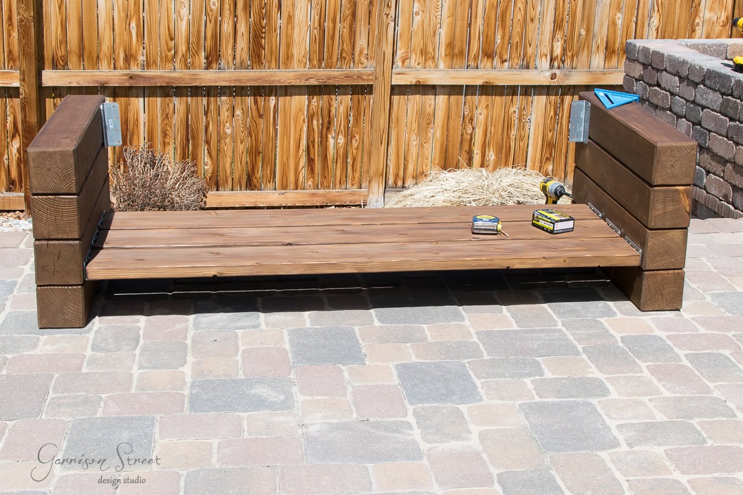 A wooden bench frame under construction on a stone patio. Two boxes of screws and a measuring square are on the seat. A wooden fence and stone wall are in the background.