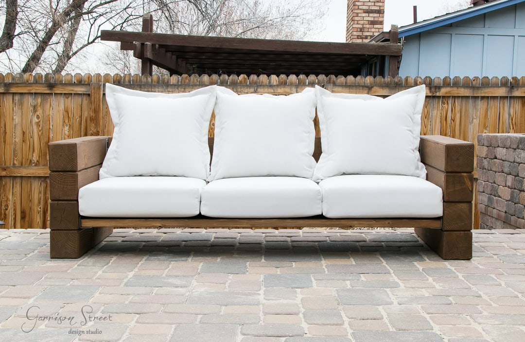 Wooden outdoor sofa with white cushions on a stone patio. The sofa is positioned in front of a wooden fence, with a pergola and brick structure visible in the background.