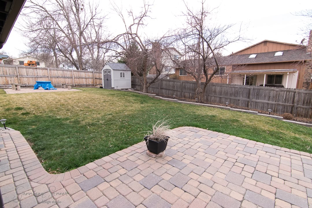 A backyard with a stone patio, a grassy lawn, and a wooden fence. There is a small garden shed in the corner and leafless trees are scattered around the yard. A planter with dried plants sits on the patio. Houses are visible beyond the fence.