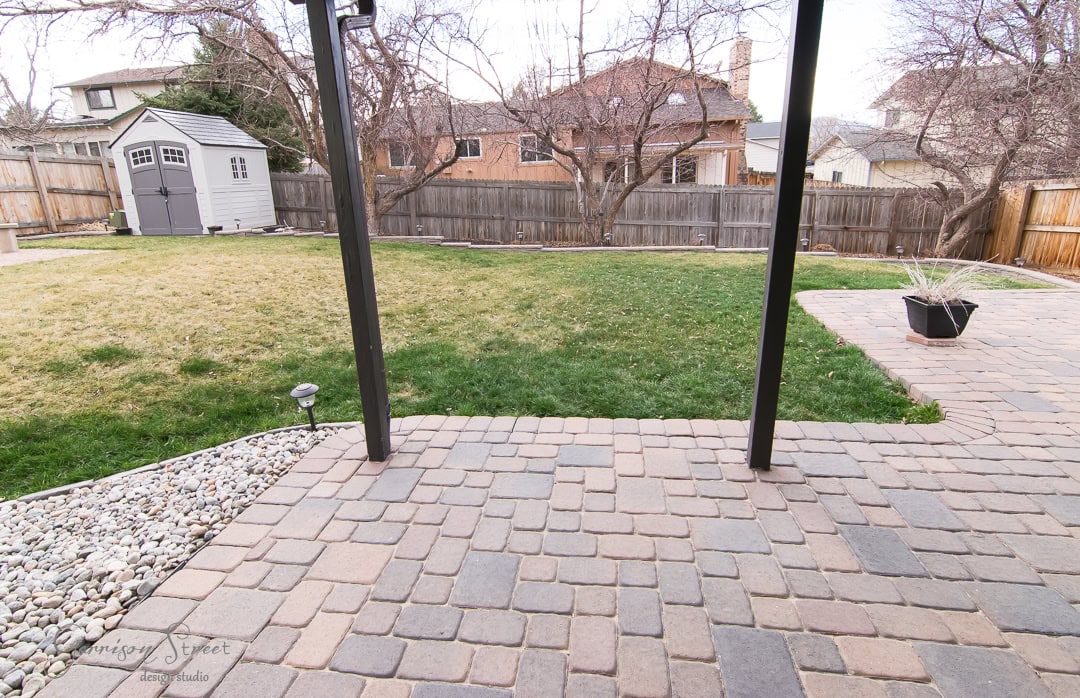 A backyard with a stone patio and partial grass lawn. A wooden shed is in the back left corner, and trees with bare branches are around the yard. A potted plant is on the patio, and a wooden fence encloses the area.