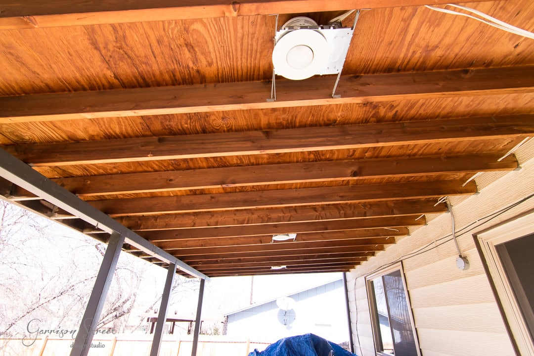 A wooden patio ceiling with visible beams, an installed round exhaust fan, and outdoor lights. The patio is attached to a house with light siding and overlooks a fenced yard with trees. A covered object sits in the corner.