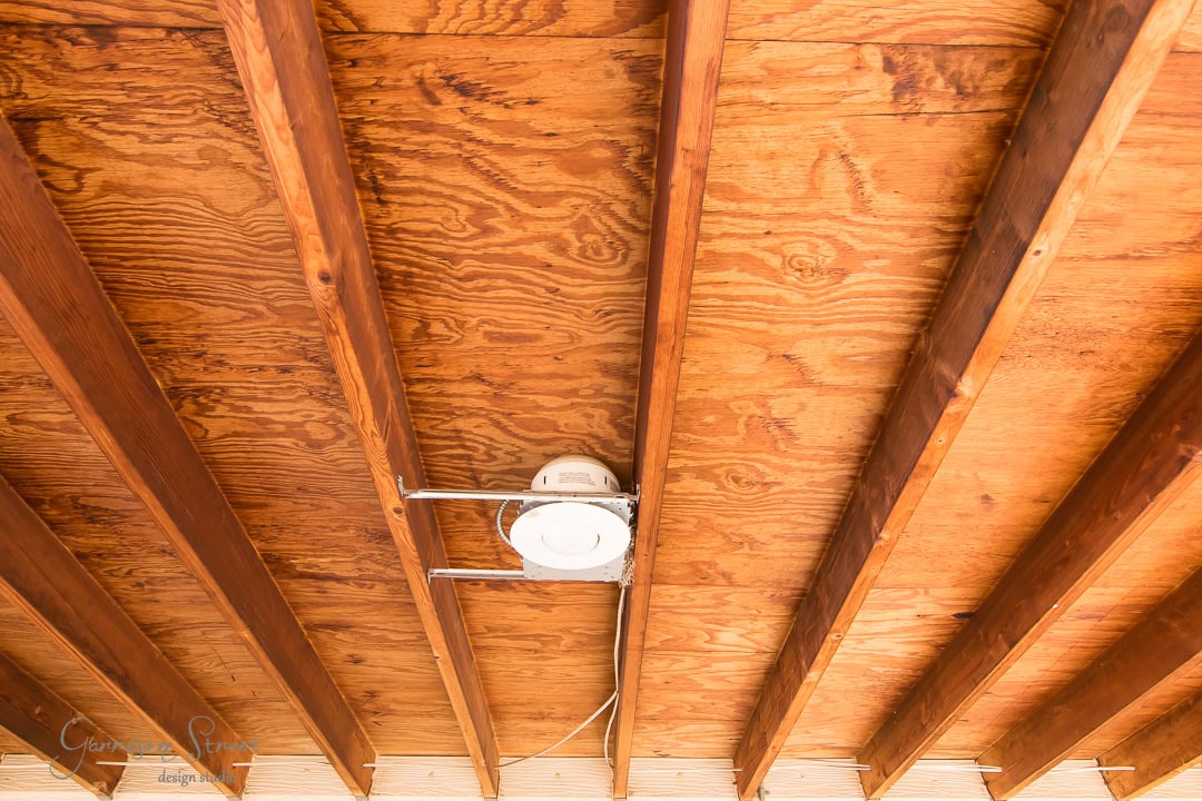 The image shows a wooden ceiling with exposed beams. A single recessed light fixture is installed in the center. The wood has a natural finish, showcasing its grain and texture.