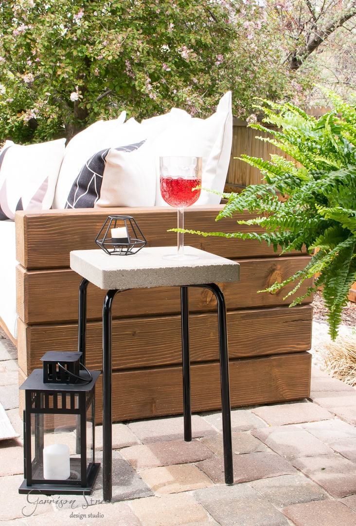 A cozy patio features a modern cement and black metal stool holding a glass of red beverage and a small candle holder. Beside it, a black lantern with a white candle adds charm. A wooden bench, adorned with cushions, completes this inviting backyard scene.