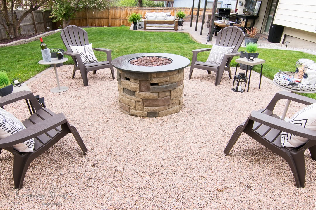 A cozy outdoor patio in the backyard features a stone fire pit surrounded by four brown Adirondack chairs on a gravel surface. Tables hold decor and a beverage, while a grassy yard and wooden fence provide a charming backdrop.