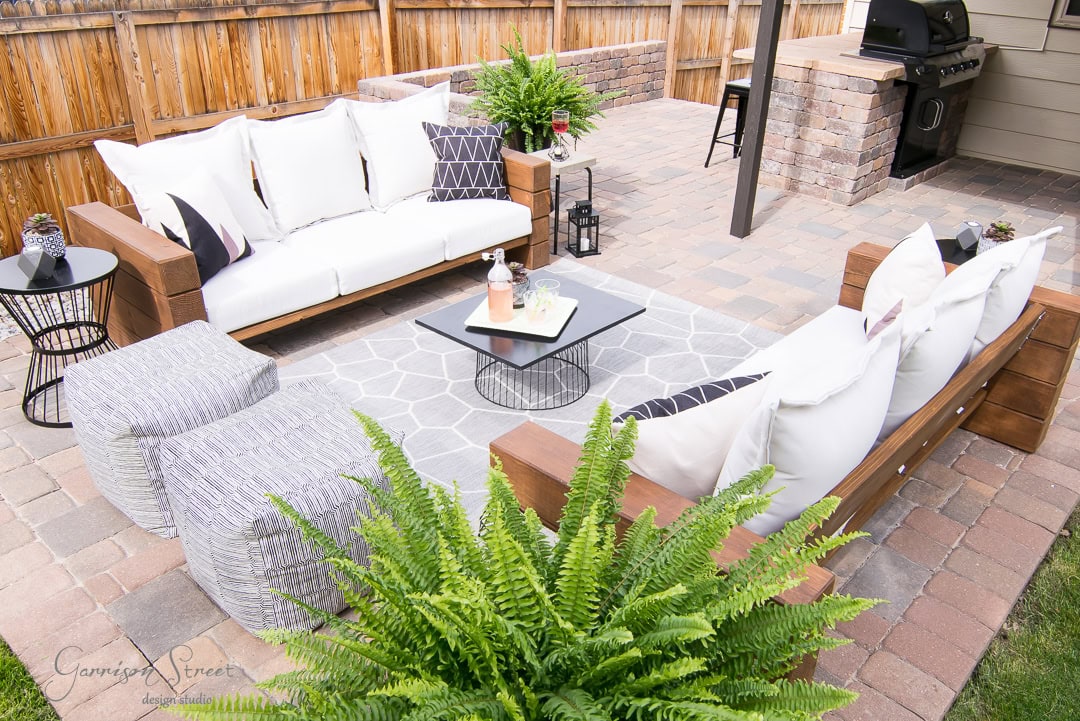 The backyard patio features wooden sofas with white cushions, a gray geometric rug, and a sleek black coffee table. Fern plants add a touch of greenery, while the brick barbecue grill stands ready for use. The stone-paved area is charmingly enclosed by a wooden fence.