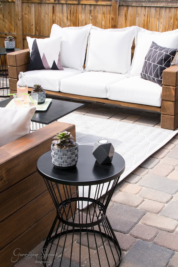 In the backyard, the outdoor patio boasts a wooden couch with white cushions and geometric pillows. A metal side table holds decorative items, while a geometric rug rests on the stone-paved floor. All is elegantly encased by a wooden privacy fence.
