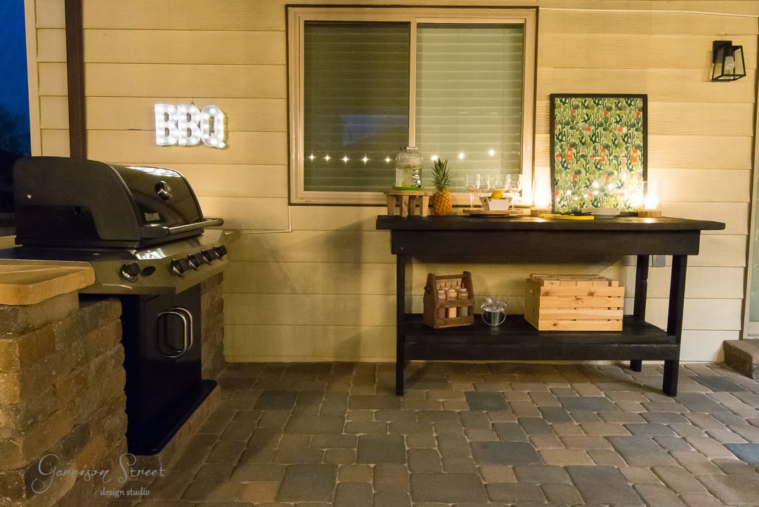 A charming backyard retreat features an outdoor patio with a barbecue grill set on a stone platform. A wooden table displays glassware, a drink dispenser, and a decorative frame, with a crate nestled below. The wall sparkles with an illuminated "BBQ" sign.