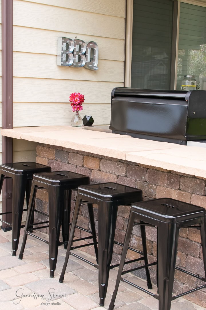 The backyard patio showcases a brick bar counter with sleek black metal stools. A vibrant pink flower arrangement in a black vase adds charm to the bar. A "BBQ" sign graces the wall, and a barbecue grill is perfectly positioned on the counter for culinary delights.