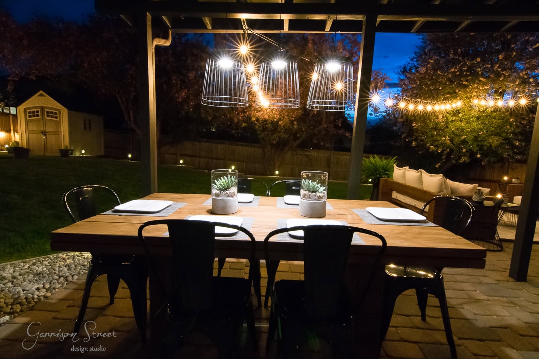 The backyard patio dining area at night features a wooden table set for four with potted plants as centerpieces. String lights and hanging lamps illuminate the space, while a cozy seating area and a shed add charm to the surroundings.