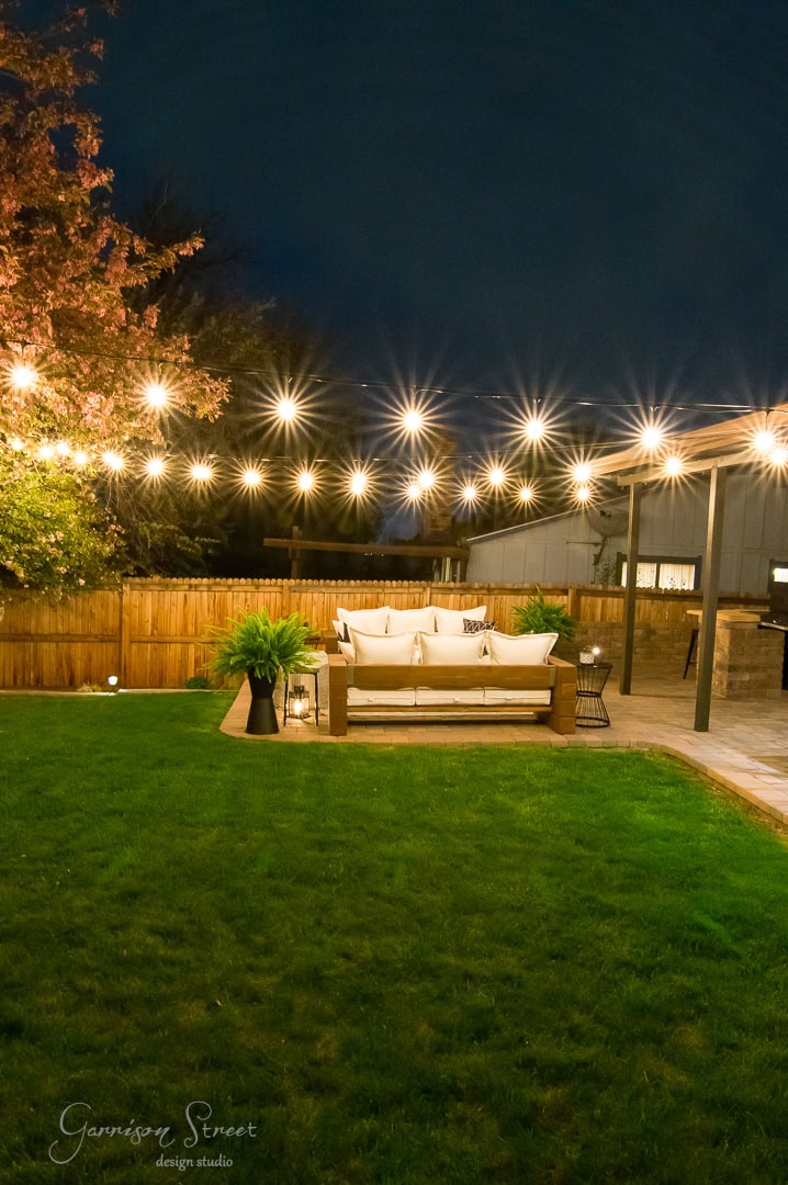A cozy backyard patio with a wooden deck features a comfortable sofa adorned with cushions. String lights hang overhead, casting a warm glow over the green lawn and adjacent wooden fence. A tree with red leaves is visible to the left, adding a touch of color to this charming outdoor space.