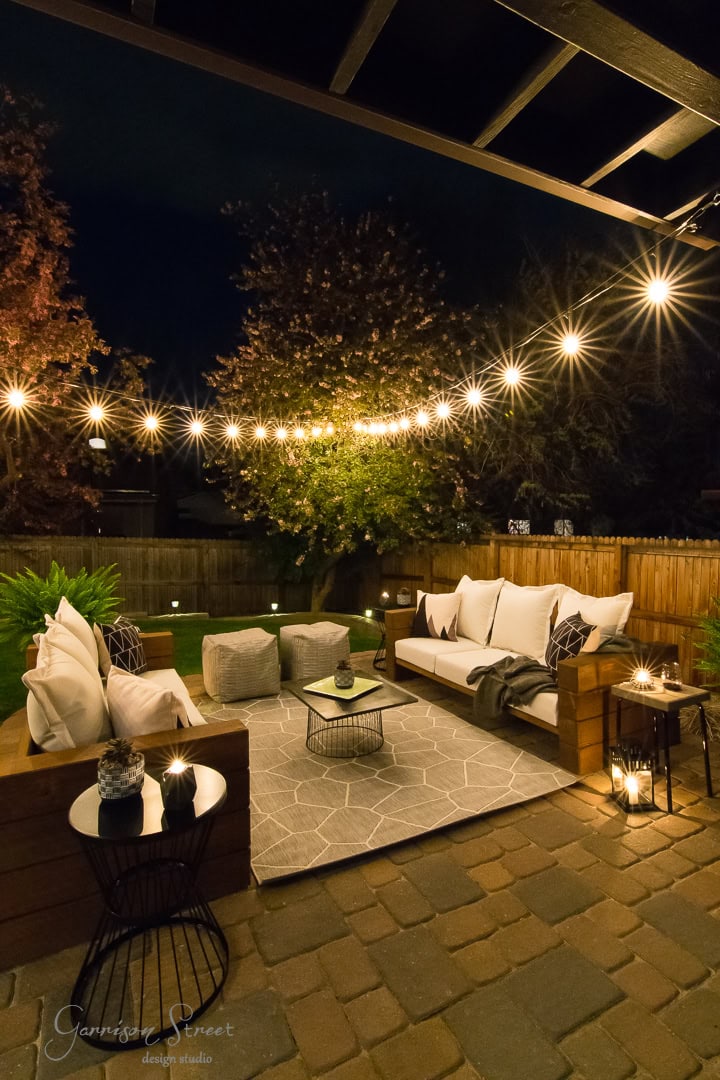 A cozy backyard patio at night with string lights hanging overhead features cushioned seating around a geometric-patterned rug. Small lights on the grass and lush greenery enhance the tranquil ambiance.