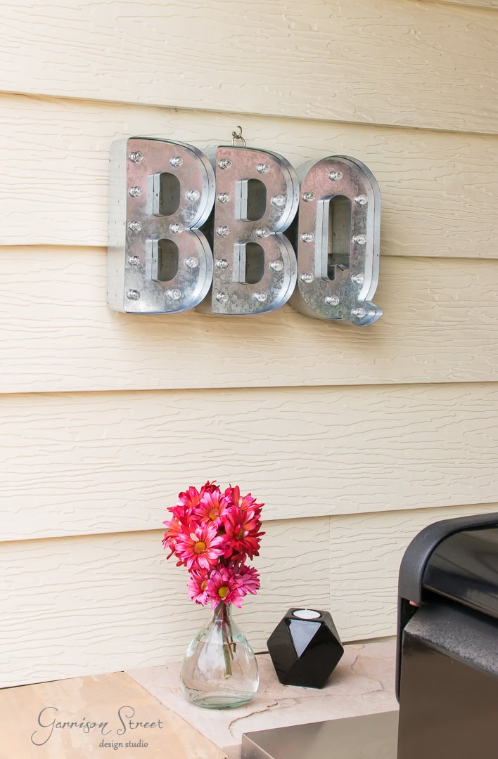 On a light-colored wall in the patio, a metallic BBQ sign with lights hangs above a small table. On the table, there's a vase with vibrant pink flowers and a black geometric candle holder. A grill is partially visible on the right.